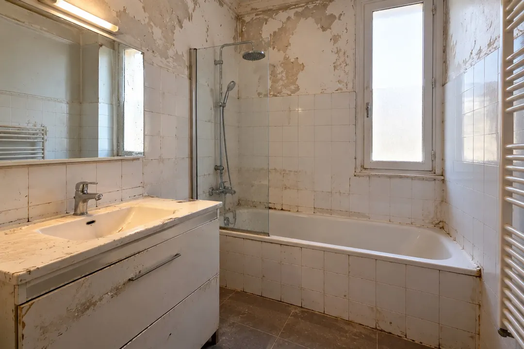 Salle de bain avant rénovation : peinture écaillée au-dessus d'une faïence ancienne encrassée, meuble vasque abîmé